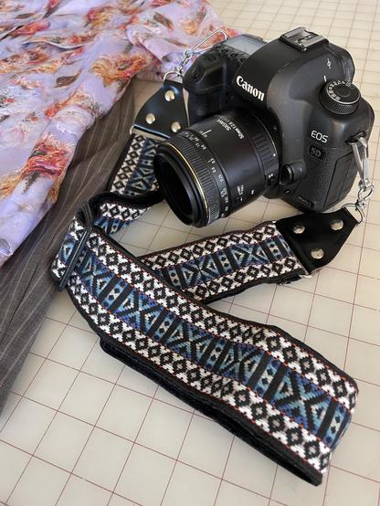 A black bodied Canon camera dominates the frame. It’s sitting on a table next to a bit of purple floral print fabric and gray fabric with pink pinstripes (a shirt and shorts respectively). Attached to the camera is a woven strap with a geometric diamond motif in blues and whites on a black background.