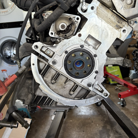 The back end of my BMW M54B30 inline-six cylinder engine, hanging from a hoist in my garage, showing off its shiny new rear main seal around the crankshaft output shaft.