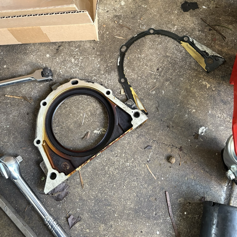 The old rear main seal assembly and gasket, removed from my BMW M54B30 inline-six cylinder engine and sitting oily-side-up on my garage floor. The mating surface between the plate around the seal is mostly clean, with some evidence of a minor possible oil leak along the bottom where the plate meets the oil pan.