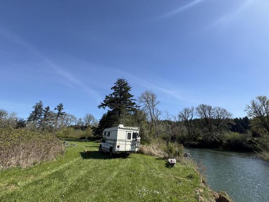 A camper is parked by a river, surrounded by grassy areas and trees under a clear blue sky.
