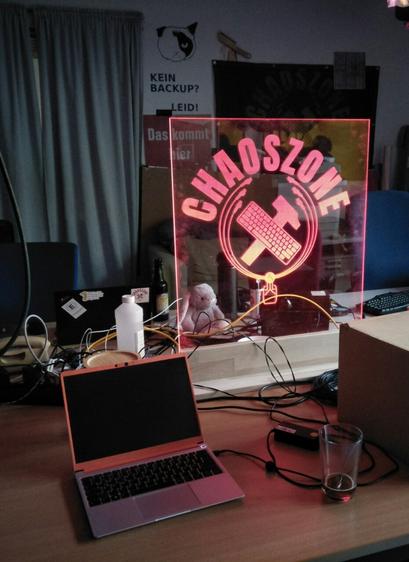 Foto von einem großen Kunststoffglas-Schild mit eingraviertem ChaosZonen Logo, rot beleuchtet. Es steht auf einem größeren Tisch auf dem noch weitere Sachen zu sehen sind. Laptops, Tastatur, Kabelage, ein Plüsch Hase, eine Flasche mit vermutlich Isopropanol, ein Karton und ein Getränk.
Im Hintergrund ein grauer Vorhang und ein Poster an der Wand mit der Aufschrift: "Kein Backup? Leid!"