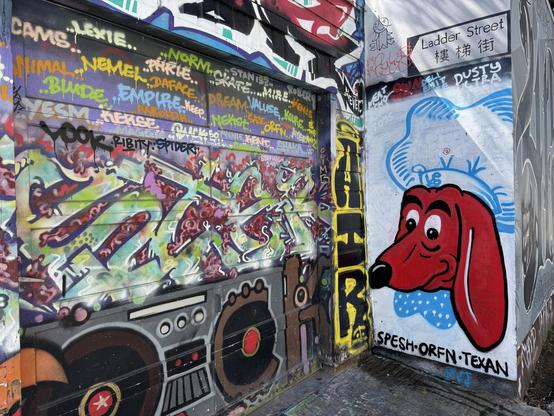 A wall in an alley that is covered with graffiti and street art. The main piece in this photo is a drawing of a red dog with a long snot and big grin. The dog is wearing a white chef's hat and white shirt. It references the logo of an old chain in the Bay Area called "Doggie Dinner".