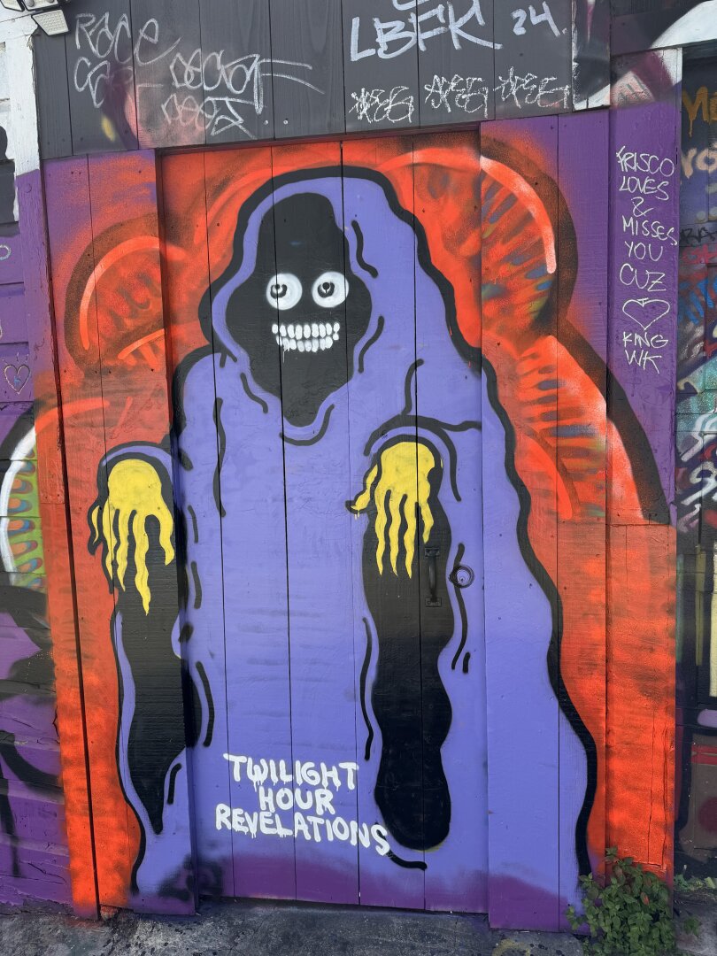Graffiti art of a wizard in a long purple robe with yellow hands poking out from the sleeves and a mischievous grin on the wizard's face. This art is drawn in an alley that is covered with graffiti and street art.