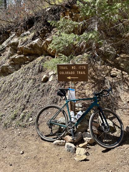 A bicycle is leaning against a sign for Trail No. 1776, the Colorado Trail. The surrounding area features rocky terrain and sparse vegetation. Two water bottles are mounted on the bike, and a notice sheet is attached to a nearby post.