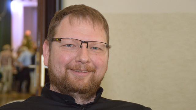 Porträt von Mitja Fritsch. Ein lächelnder Mann mittleren Alters mit roten Haaren und Bart. Er trägt eine schwarze, hochgeschlossene Kleidung und eine Brille. Der Hintergrund ist unscharf, zeigt aber einen Raum mit weiteren Personen.