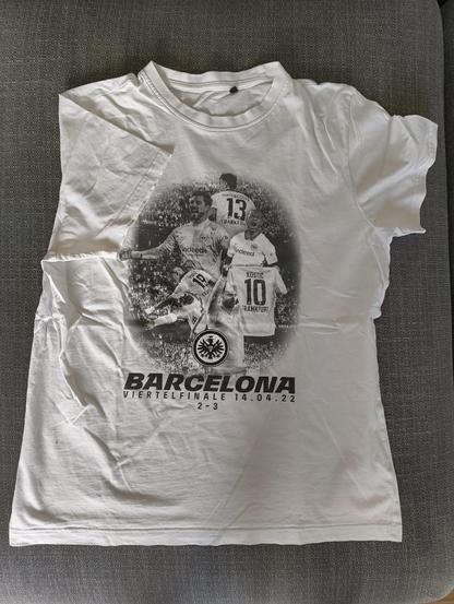Das Bild zeigt ein weißes T-Shirt, das auf einem grauen Sofa ausgebreitet liegt. Auf dem T-Shirt befindet sich ein Schwarz-Weiß-Druck, der drei Fußballspieler zeigt, die in Aktion sind. Die Spieler tragen Trikots mit den Nummern 13, 19 und 10, und die Namen "BORRE" und "KOSTIC" sowie "HINTEREGGER" sind auf den Rücken der Trikots zu sehen. Der Druck enthält auch den Text "BARCELONA VIERTELFINALE 14.04.22 2 - 3" und das Logo des Fußballvereins Eintracht Frankfurt. Der Druck erinnert an ein Fußballspiel zwischen FC Barcelona und Eintracht Frankfurt am 14. April 2022, bei dem Eintracht Frankfurt mit 3:2 gewann.