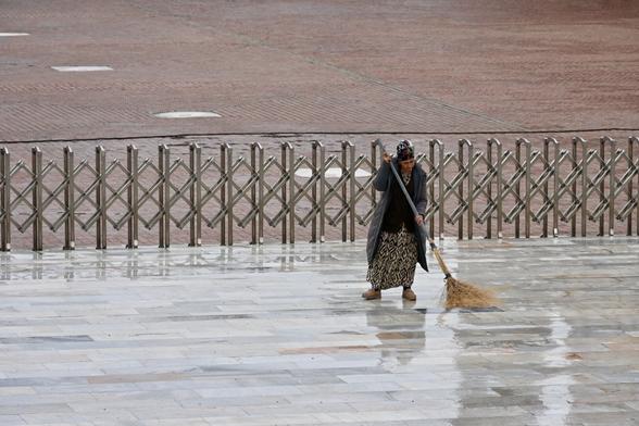 A woman sweeping a rainy square with a broom. Some washed out shapes of surrounding buildings are reflected in the wet surface.