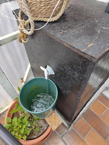 Auf einem Balkon läuft aus einem Hahn an  einer Holzkiste Wasser in einen Eimer. In der Kiste wird Regenwasser von der Dachrinne gesammelt.