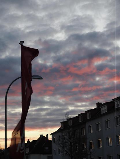 The image features a dramatic sky with clouds illuminated by sunset colors, primarily shades of pink and orange. In the foreground, there's a red flag attached to a pole, and a streetlight is visible next to it. Residential buildings are in the background, silhouetted against the colorful sky.