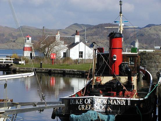 Crinan Harbour. The image shows a tug-type boat with the name Duke of Normandy II on its stern on the right-hand side of the frame. It has a black hull and red funnel. The area of water it is in is surrounded by a low stone edging with grass beyond. In the background is a white cottage and a small red and white striped lighthouse. Beyond is open water and mountains in the distance.