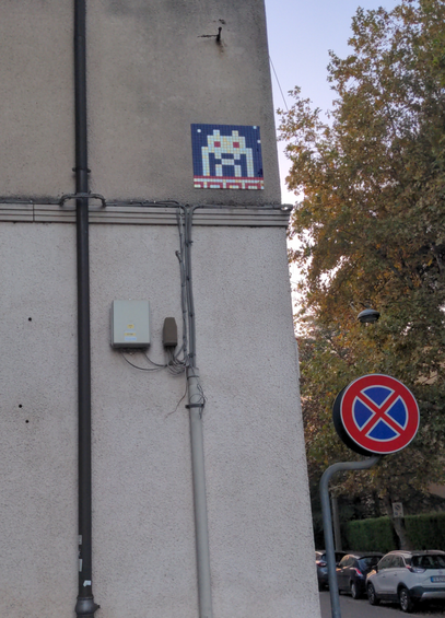 À un coin de rue gris, en hauteur, un petit invader en mosaïque blanche sur fond bleu, les yeux rouges, avec un motif rouge sur blanc en frise du bas.
