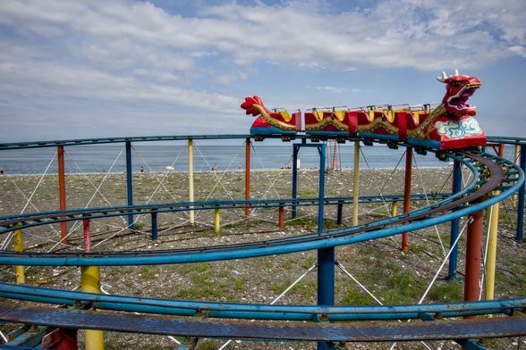 Eine bunt lackierte, aber ziemlich verrostete Miniacrhterbahn für Kinder. Die Wagen bilden zusammen einen Drache mit zwei Köpfen. Im Hintergrund sieht man einen Kiesstrand und das Meer.