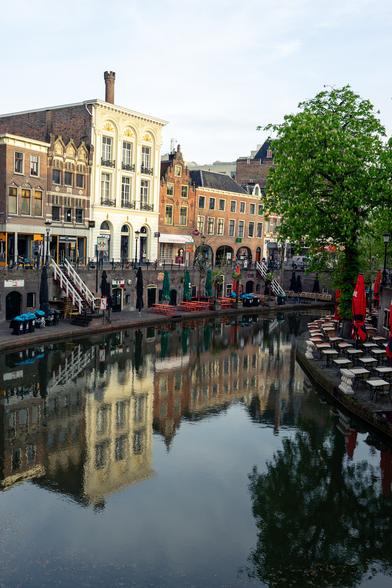 The image depicts a picturesque canal scene in a European city, likely in the Netherlands. The foreground features a calm canal with clear reflections of the surrounding buildings and trees. On the left side, there are several historic buildings with distinct architectural styles, including brick facades and ornate window frames. The buildings have a mix of colors, with a prominent white building in the center. The right side of the canal shows a tree with lush green leaves and outdoor seating with red umbrellas and tables, suggesting a café or restaurant area. The sky is partly cloudy, and the overall atmosphere is serene and inviting.

Provided by @altbot, generated privately and locally using Ovis2-8B

🌱 Energy used: 0.161 Wh