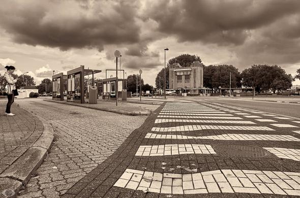 Black and white image of bus stop at left bank, Antwerp. 1/3 of the image (right bottom) takes up space in the form of zebra crossing that the busses have to cross when they leave. As a result, this is crooked as the wheels have bent the stones. Completely at the left is a woman, presumably waiting for the bus and checking her phone. Lots of dark clouds that make a threatening sky.