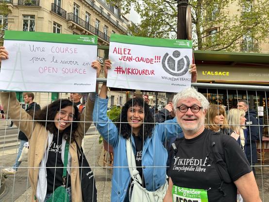 Jean Claude Balès entouré de Sana et Alejandra lors du Marathon de paris. Sana et Alejandra tiennent des pancartes d'encouragement qui encourage Jean-Claude et valorise les projets WIkimédia France