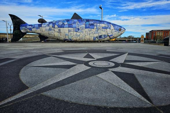 A wide shot captures a large, mosaic-tiled fish sculpture on a paved area featuring a compass rose design. The sky is blue with scattered clouds.