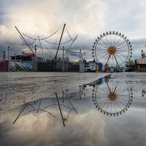 Das Bild zeigt einen Jahrmarkt im Aufbau. Im Vordergrund liegt eine große Pfütze, die eine klare Spiegelung der Umgebung zeigt. Im Hintergrund thront ein imposantes Riesenrad mit gelben und roten Streben sowie schwarzen Gondeln. Links daneben ragen hohe Masten auf, vermutlich Teil eines Fahrgeschäfts mit gespannten Seilen. Mehrere Stände und Buden mit bunten Schildern und Bannern, sowie geparkte Autos sind ebenfalls erkennbar. Der Himmel ist bewölkt, was dem gesamten Bild eine leicht düstere, aber faszinierende Atmosphäre verleiht.