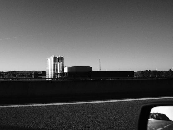 Monochrome, medium-shot photograph. The foreground showcases highway asphalt with a horizontal lane marking line, leading the eye to a continuous barrier wall slightly out of focus. Behind this barrier, the primary subject is an industrial complex composed of several interconnected cylindrical silos with bright reflective surfaces and a long, dark-colored rectangular building. Distant windmills and a telecommunications tower are faintly visible on the horizon under a vast, clear sky. Lower right corner shows a small portion of a car's side mirror, suggesting the photograph was taken from inside a vehicle in motion. The grayscale palette emphasizes the interplay of light and shadow across the scene.