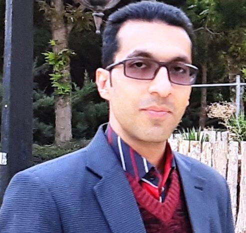 A man with dark, relatively short hair and slightly tinted glasses looks at the camera. He is wearing a red striped jumper and shirt with a dark cord jacket. There are trees in the background.