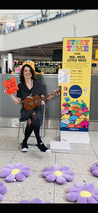 Me in terminal 2 with a mr strong plushie and a ukulele. There is a big sign Saying story time behind me