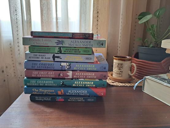 A stack of books with a mug and a plant in a table in front of a window curtain