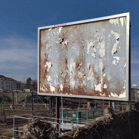 Un ancien panneau publicitaire à l'abandon devant des voie de chemin de fer. La rouille est apparente. Le mot "Avenir", est disposé en haut, telle une marque.
