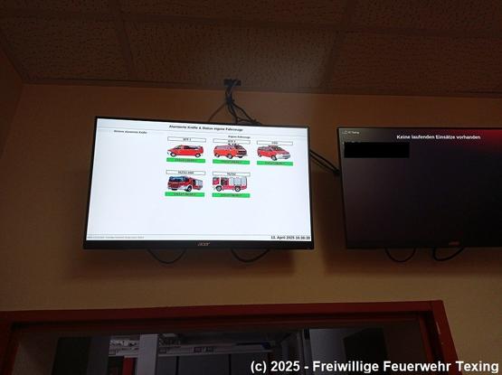 Zwei Cmoputermonitore an der Wand befestigt. Die Monitore zeigen Einsatzdaten. Darunter eine rote Brandschutztüre.