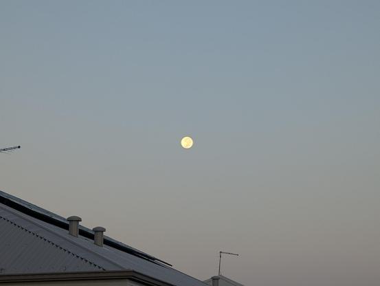 This image depicts a serene scene of a full moon in the sky, positioned above the rooftops of residential buildings. The moon appears bright and prominent against a clear, light blue sky, suggesting that the photo was taken either during dusk or dawn. The rooftops are equipped with chimneys and antennas, indicating a suburban setting. The overall atmosphere of the image is calm and peaceful, capturing a quiet moment in a residential area.