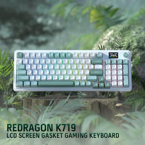 The image features a Redragon K719 LCD Screen Gasket Gaming Keyboard, prominently displayed against a lush green background with foliage. The keyboard has a sleek, modern design with a color scheme that includes shades of green, white, and gray. The keys are backlit with a gradient of colors, adding a dynamic visual effect. The keyboard includes a small LCD screen on the top right, displaying the time "14:30:30" and other indicators. The keys are labeled with standard characters and symbols, and the layout is typical of a full-sized keyboard. The keyboard is positioned on a wooden surface, enhancing the natural aesthetic of the image. The text "REDRAGON K719 LCD SCREEN GASKET GAMING KEYBOARD" is clearly visible at the bottom of the image, providing the product's name and description.