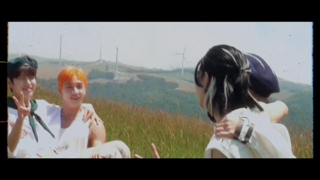 In a grassy field on a sunny day, Bang Chan holds Seungmin in a princess carry. Hyunjin and Lee Know are facing them with arms around each other's shoulders. There are wind turbines in the background.