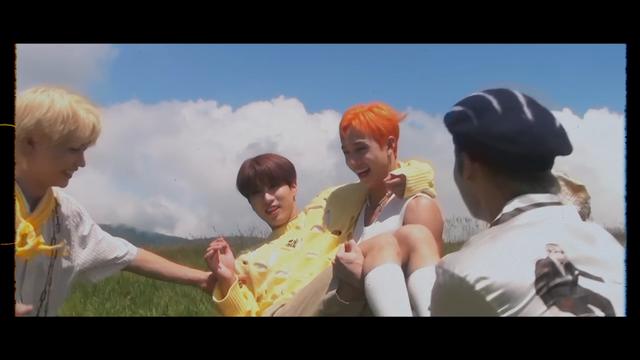 In a grassy field on a sunny day, Bang Chan holds Han in a princess carry, while Felix and Lee Know watch.