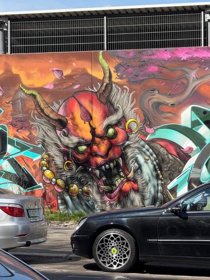 A mural depicting a Japanese Oni, a red skinned demon like creature, bald, with a grey beard and side burns, green eyes, his teeth and tusks in various states of white, with a few teeth being gold, and he's wearing a gold necklace and earring.