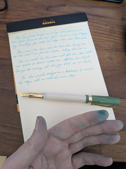 A picture of my hand stained with turquoise ink in front of a blank-paper Rhodia pad.  On it in bright, light blue are the lyrics to Slow Parade by Broken Records, written in my scrawled roundhand style.  The Kaweco Student lies on top with its cap off and on the pen's back end.  Its nib and grip are gold, its body ivory, the cap a pastel green, with gold trim and clip.