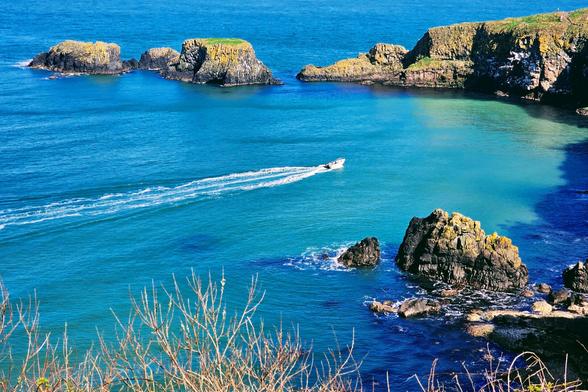 A high-angle shot of a turquoise bay with rocky cliffs, small islands, and a white boat leaving a trail in the water.