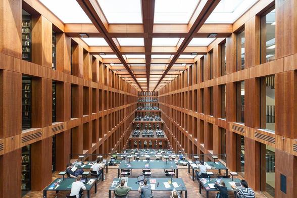 A large library interior with grid-like ceiling and wall pattern. It is a long rectangular space with a series of descending terraces, filled with large work tables on which people are working.