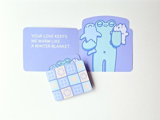 A picture of two cards, the big one of top shows an opened purple greeting card with the text on the left: Your love keeps me warm like a winter blanket. On the right there's an illustration of a frog cuddling a blue shark and a white bear plush. Below there's a smaller closed card with just the heads of frog and friends peeking out under a blue, yellow and purple patchwork blanket.