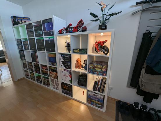 Two Kallax shelves filled with stuff, image taken from the right at an angle. Most of the compartments hold boardgames, a few have Lego models or plushies, and very few are still empty. On top of the near, shorter shelf, there's a Lego Ferrari and a Lego plant. On top of the taller shelf, all the way in the back and in the corner, there's a game box of "City Of Angels" boardgame. Behind the taller shelf on the left and further back, green lights shine through from behind the shelf. The front one has warm white spots in every other compartment.