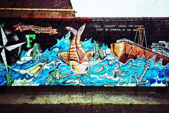 A vibrant and detailed nautical-themed mural painted on a dark brick wall. The artwork features swirling blue and white waves dominating the lower half, suggesting a turbulent sea. Above the waves, a large, orange fish with prominent fins is seemingly caught in the current. To the right, a portion of a weathered brown and grey ship is visible, with its tall mast extending upwards. An anchor is drawn near the ship, firmly planted in the imagined seabed.
Above the central fish, a text overlay reads "YOU CANNOT CROSS THE OCEAN JUST BY STARING AT THE SEA," rendered in simple white lettering.