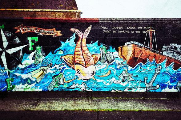 A vibrant and detailed nautical-themed mural painted on a dark brick wall. The artwork features swirling blue and white waves dominating the lower half, suggesting a turbulent sea. Above the waves, a large, orange fish with prominent fins is seemingly caught in the current. To the right, a portion of a weathered brown and grey ship is visible, with its tall mast extending upwards. An anchor is drawn near the ship, firmly planted in the imagined seabed.
Above the central fish, a text overlay reads "YOU CANNOT CROSS THE OCEAN JUST BY STARING AT THE SEA," rendered in simple white lettering.