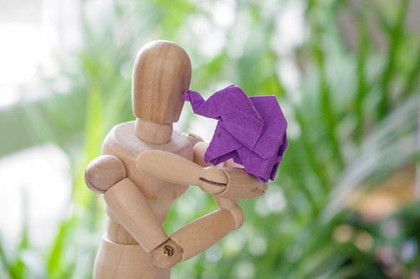 Nahaufnahme eines kleinen lila Origami-Elefanten, der auf den armen einer Gliederpuppe (zum Zeichnen) steht und sie mit seinem Rüssel quasi küsst.