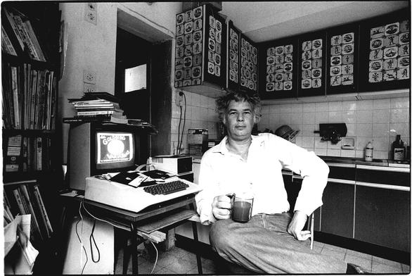 Yaakov Kirschen sits in his kitchen holding a drink. Beside him is an Apple II computer on a desk. The computer monitor shows the title screen of a game called "The Goldberg Variations."