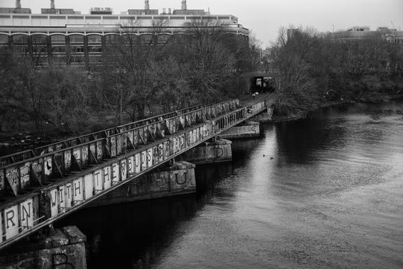 A graffiti-covered railroad bridge stretches over a calm river, with bare trees lining the banks. In the background, industrial buildings are visible. The scene is presented in black and white, creating a moody atmosphere.
