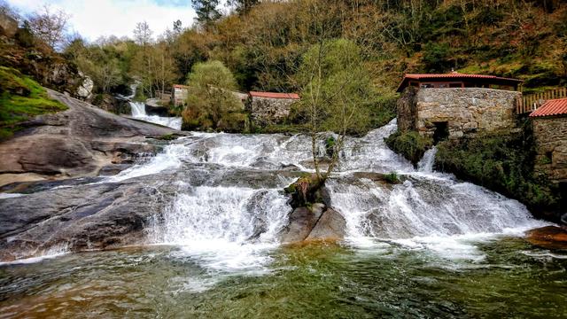 A wide shot captures a cascading waterfall flowing over smooth, dark rocks, with several old stone mill buildings featuring red-tiled roofs nestled amongst green trees on the banks. The water is foamy white as it rushes downwards, creating a dynamic contrast with the still, mossy rocks and the solid structures.