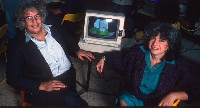Yaakov Kirschen and Sali Ariel are seated, smiling at the camera. Between them is a Commodore Amiga displaying a large caricature  of Bach from Kirschen's "Magic Harp" software.