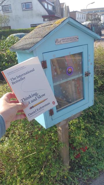 Small blue wooden mini library on a pole, with a glass door. On left, a hand holds a book titled "thinking, fast and slow" by Daniel Kahneman