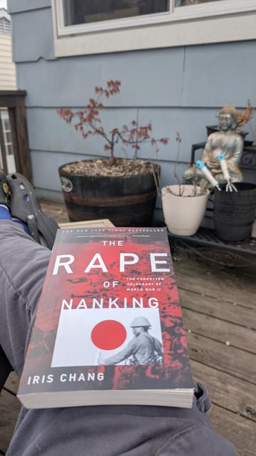 A person sits outside on a wooden deck with their legs stretched out, holding a paperback copy of The Rape of Nanking by Iris Chang. The book's cover features a soldier and the red circle of the Japanese flag. In the background is a peaceful garden setup: a potted Japanese maple tree with red leaves, several other potted plants, and a Buddha statue. The Buddha has two blue-handled gardening tools resting in front of it. The scene contrasts the serenity of the setting with the serious historical subject