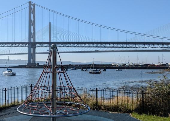 In the foreground is a cone climber for children, made of rope netting suspended between a central mast and a wide rotating circle. The climber's overall shape is roughly triangular. Behind it two bridges span the water – a suspension bridge, whose deck is supported by a system of triangular trusses, and a cable-stayed bridge, whose sail-like cables form numerous triangles. On the water and docked nearby are a dozen or so sailboats with triangular masts and rigging.