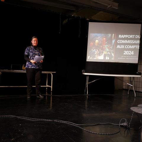 À gauche une femme lit ses notes, à droite une projection avec noté "rapport du commissaire aux comptes 2024" dans une salle sombre.