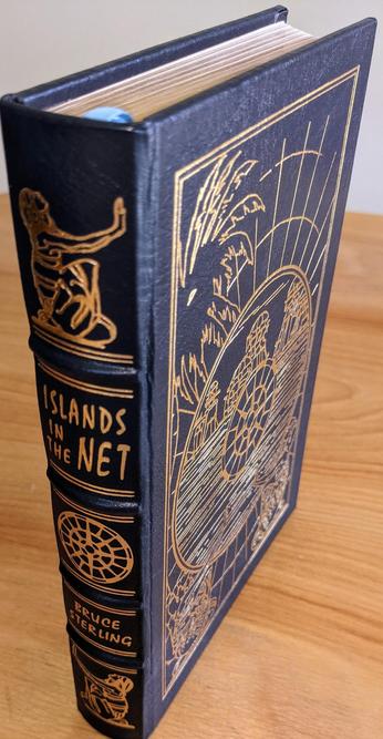 Stunning Easton Press limited collector's edition of Islands in the Net by Bruce Sterling from the prestigious Masterpieces of Science Fiction series. This edition is bound in full genuine leather, dyed a deep dark navy blue (almost black), with elaborate 22kt gold embossing on both cover and spine. The front cover features original artwork by Kent Bash, rendered in 22k gold leaf, with a network-like schematic surrounding an island paradise, encapsulated in a stylized grid structure. Introduction by James Gunn
 - 
The spine is hubbed and hand-tooled, featuring a geometric satellite-like emblem and two intricate vignettes of seated human figures using terminals, evoking both Sterling's cyberpunk roots and the narrative's geopolitical techno-surveillance themes. The title and author's name are boldly stamped in 22k gold, perfectly aligned with the edition's archival standards
 - 
Printed on acid-neutral archival paper, the pages are 22k gold gilded on all edges, giving the book a brilliant reflective sheen. Smyth sewing and concealed muslin joints ensure a lifetime of structural integrity. This copy is unread, from a single-owner private collection, and has been stored exclusively in dust-free, climate-controlled bookcases, preserved in pristine, as-new condition. The bookplate inside the front cover remains untouched. A true artifact for collectors, showcasing craftsmanship and literary significance in equal measure