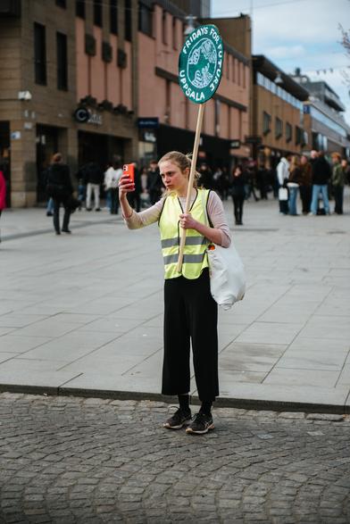 A protester with green safety jacket holding a "Fridays for Future" placard and taking a photo on mobile just when the march started.